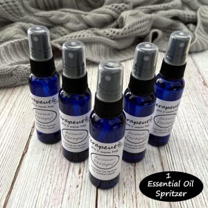 May include: Five blue glass bottles with black spray tops and white labels that read "Therapeutic, Give a warm hug, Aroma-Ferapy! Essential Oil Spritzer for comfort packs". The bottles are arranged in a circle on a wooden surface. The text "1 Essential Oil Spritzer" is written in white on a black circle below the bottles.