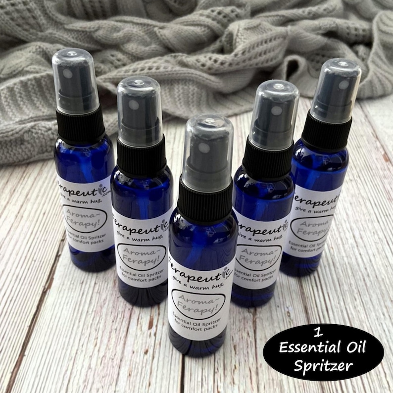 May include: Five blue glass bottles with black spray tops and white labels that read "Therapeutic, Give a warm hug, Aroma-Ferapy! Essential Oil Spritzer for comfort packs". The bottles are arranged in a circle on a wooden surface. The text "1 Essential Oil Spritzer" is written in white on a black circle below the bottles.