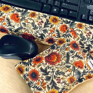 May include: A rectangular fabric wrist rest and a matching mouse pad featuring a cream background with a dense floral pattern of orange, red, and yellow flowers interspersed with dark green foliage. A black computer keyboard and mouse are present.