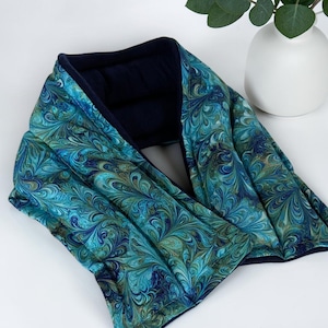 A hot or cold neck wrap featuring a vibrant turquoise blue marbled fabric and navy interior lining.  The wrap has a soft, quilted texture and a small white label with text.