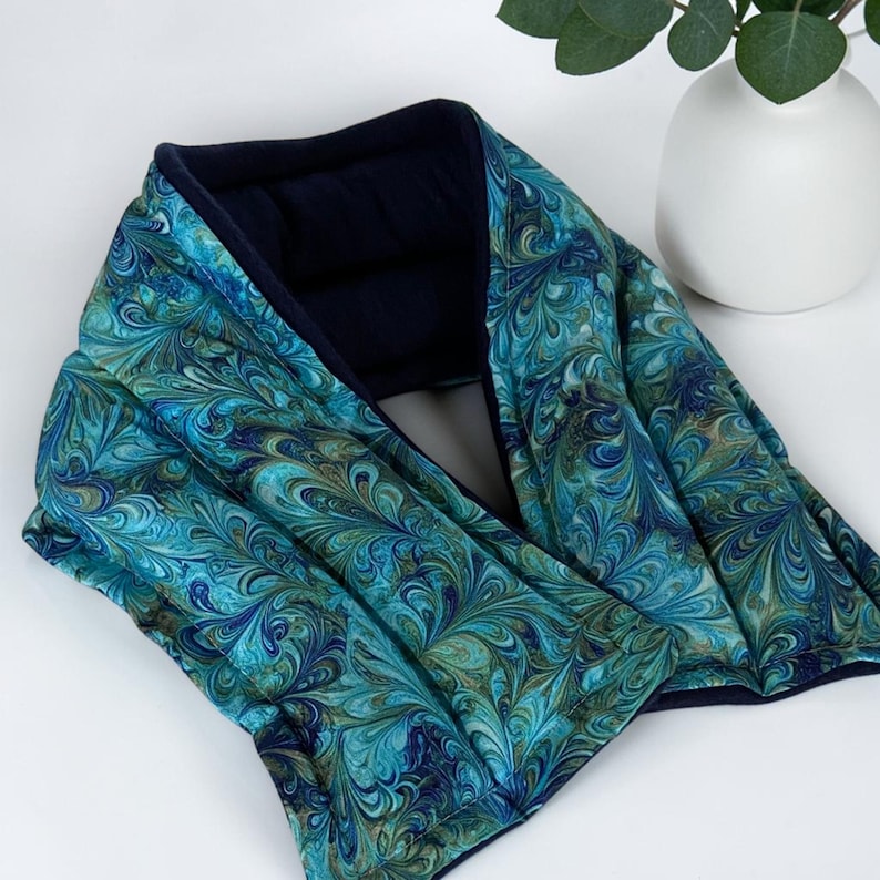A hot or cold neck wrap featuring a vibrant turquoise blue marbled fabric and navy interior lining.  The wrap has a soft, quilted texture and a small white label with text.