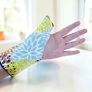 May include: A wrist and thumb support brace featuring a vibrant floral pattern with overlapping petals in shades of light blue, bright yellow, and burnt orange against a white background, with a black interior lining visible at the edges.