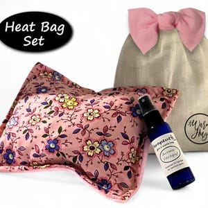 K&ouml;nnte beinhalten: Ein W&auml;rmekissen-Set bestehend aus einem rechteckigen rosa Kissen mit buntem Blumenmuster, einem beigen Kordelzugbeutel mit rosa Schleife und einer kleinen blauen Spr&uuml;hflasche mit der Aufschrift "Terapeutic Aroma-Ferapy" und "A Warm Hug".