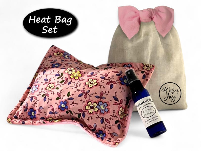 K&ouml;nnte beinhalten: Ein W&auml;rmekissen-Set bestehend aus einem rechteckigen rosa Kissen mit buntem Blumenmuster, einem beigen Kordelzugbeutel mit rosa Schleife und einer kleinen blauen Spr&uuml;hflasche mit der Aufschrift "Terapeutic Aroma-Ferapy" und "A Warm Hug".