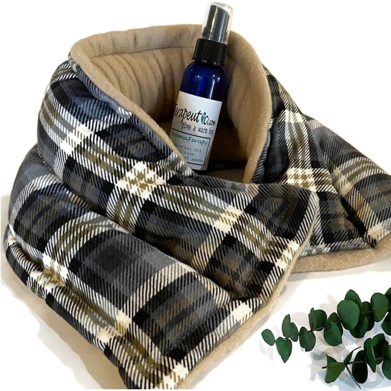 A gray, black, and white plaid neck wrap with a tan fleece lining. A small blue bottle with a white label that reads 'Therapetuc Calm give a warm hug Aromatherapy Essential Oil Diffuser Heat Packs' is inside the neck wrap.
