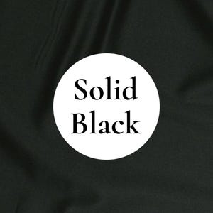 May include: A solid black fabric background featuring a centered white circle containing the black text Solid Black. The fabric has a smooth, matte texture with subtle folds and shadows that create a soft, uniform appearance across the surface.