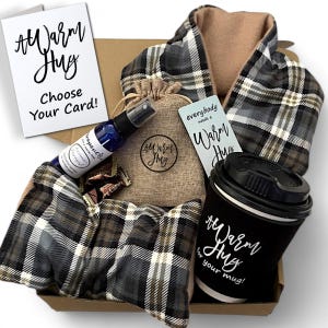 May include: A gift box filled with self-care items, including a plaid scarf, a small burlap bag with a brown label that says "#Warm Hug", a bottle of aromatherapy spray, a card that says "everybody needs a warm hug", a mug with a black sleeve that says "#Warm Hug for your mug!" and a few chocolate candies.