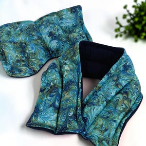 A long, rectangular neck wrap featuring blue marbled fabric in shades of turquoise, green and blue. There is a sectioned eye pillow in the image. The interior lining is a solid, textured navy fabric. A small white tag with the text "Ferapeutic" is attached to the edge.