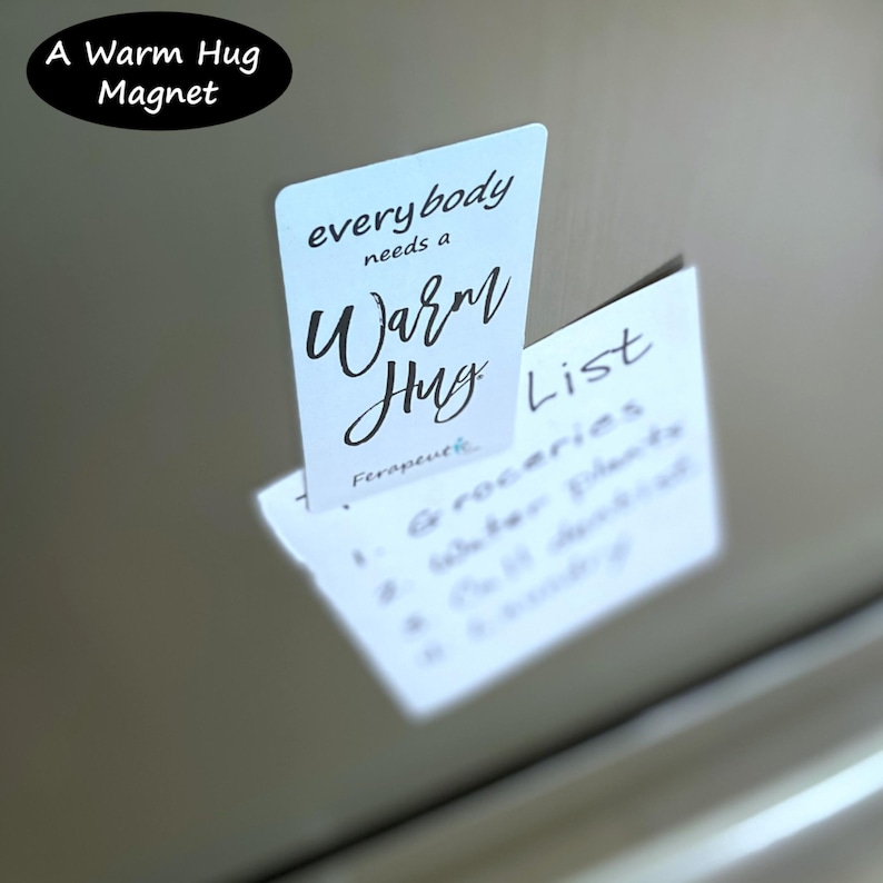 May include: White refrigerator magnet with the text "everybody needs a warm hug" in black cursive font. The magnet is attached to a white refrigerator with a handwritten grocery list.