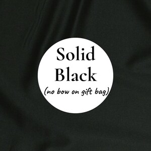 K&ouml;nnte beinhalten: Ein einfarbiger schwarzer Stoffhintergrund mit einer wei&szlig;en kreisf&ouml;rmigen Grafik in der Mitte. Der Text im Kreis lautet Solid Black und darunter in kleinerer Schrift (no bow on gift bag). Der Stoff weist sichtbare Falten und eine matte Textur auf.