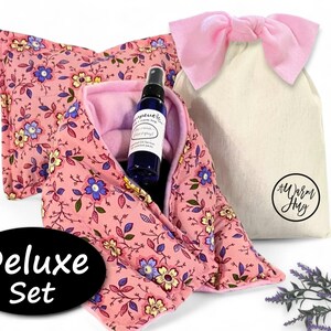 K&ouml;nnte beinhalten: Ein rosafarbenes W&auml;rmekissen aus Stoff mit Blumenmuster, eine blaue Spr&uuml;hflasche mit wei&szlig;em Etikett und ein cremefarbener Kordelzugbeutel mit rosa Schleife. Der Beutel hat ein kreisf&ouml;rmiges Logo mit dem Text A Warm Hug. Ein schwarzes Oval zeigt Deluxe Set.
