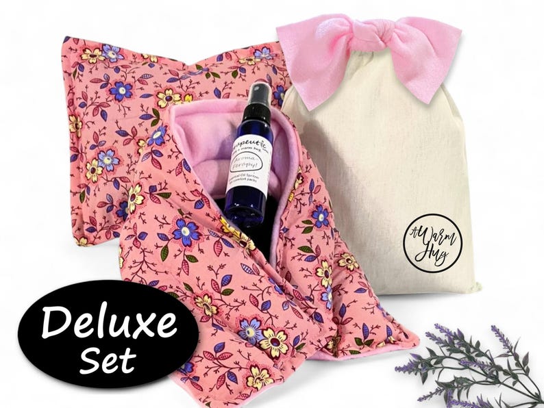 K&ouml;nnte beinhalten: Ein rosafarbenes W&auml;rmekissen aus Stoff mit Blumenmuster, eine blaue Spr&uuml;hflasche mit wei&szlig;em Etikett und ein cremefarbener Kordelzugbeutel mit rosa Schleife. Der Beutel hat ein kreisf&ouml;rmiges Logo mit dem Text A Warm Hug. Ein schwarzes Oval zeigt Deluxe Set.