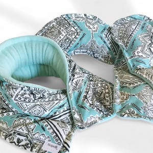 May include: A turquoise blue neck wrap with a white and black geometric pattern. The wrap is made of soft fabric and has a fleece lining. The wrap is folded in half and the ends are overlapping.