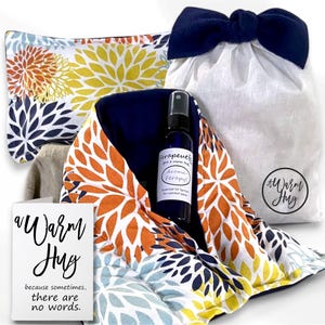 May include: A set containing a rectangular heating pad with a floral pattern in orange, yellow, and blue, a dark blue spray bottle labeled Aromatherapy, a white drawstring bag with a dark blue bow, and a card with the text A Warm Hug.