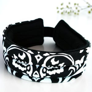 May include: A black and white headband featuring a bold, repeating damask-style pattern. The accessory has a thick, padded construction with a solid black interior lining and a flexible band design that curves into a circular shape.