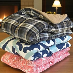 May include: A stack of four quilted pads in various patterns. The top pad has a plaid design in shades of gray, black, and white. Below it is a pad with a blue and white geometric pattern, followed by a pink paisley pad.