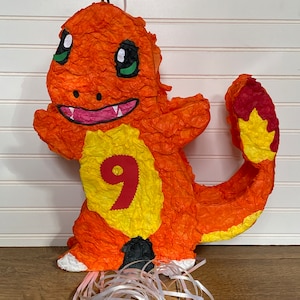 Op de afbeelding: Een oranje en gele papier-maché piñata in de vorm van een cartoonfiguur met het nummer 9 op zijn borst. De piñata heeft een zwart touwtje dat van boven hangt en witte slingers aan de onderkant.