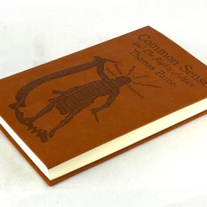 May include: A brown hardcover book titled "Common Sense and The Rights of Man" by Thomas Paine. The cover features a debossed illustration of a figure and text. The book has cream-coloured pages and a red spine.