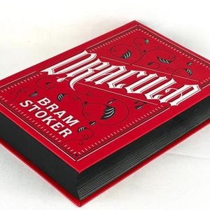 May include: A hardcover book titled Dracula by Bram Stoker with a vibrant red cover featuring white lettering and black decorative scrollwork. The book has black page edges and is positioned at an angle to show the front cover and the black spine.