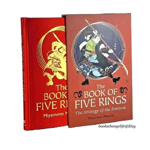 K&ouml;nnte beinhalten: Zwei rote Hardcover-B&uuml;cher mit goldener Beschriftung. Das Buch links tr&auml;gt den Titel "The Book of Five Rings" mit dem Namen des Autors "Miyamoto Musashi" darunter. Das Buch rechts tr&auml;gt den Titel "The Book of Five Rings: The Strategy of the Samurai" mit dem Namen des Autors "Miyamoto Musashi" darunter. Beide B&uuml;cher zeigen Illustrationen von Samurai-Kriegern auf dem Cover.