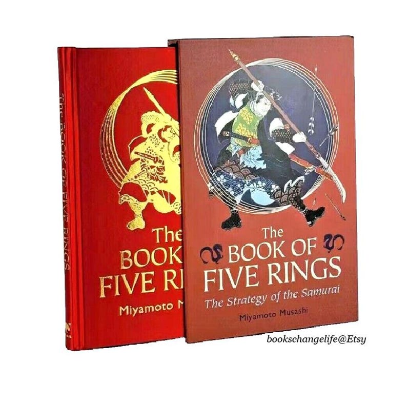 K&ouml;nnte beinhalten: Zwei rote Hardcover-B&uuml;cher mit goldener Beschriftung. Das Buch links tr&auml;gt den Titel "The Book of Five Rings" mit dem Namen des Autors "Miyamoto Musashi" darunter. Das Buch rechts tr&auml;gt den Titel "The Book of Five Rings: The Strategy of the Samurai" mit dem Namen des Autors "Miyamoto Musashi" darunter. Beide B&uuml;cher zeigen Illustrationen von Samurai-Kriegern auf dem Cover.
