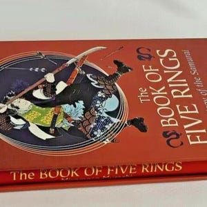 K&ouml;nnte beinhalten: Ein rotes Hardcover-Buch mit dem Titel "Das Buch der F&uuml;nf Ringe: Die Strategie des Samurai" von Miyamoto Musashi. Der Umschlag zeigt eine Illustration eines Samurai-Kriegers in Aktion.