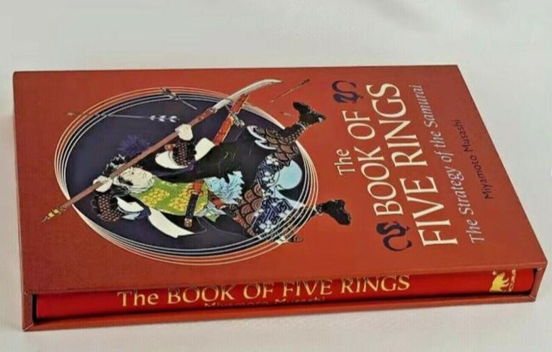 K&ouml;nnte beinhalten: Ein rotes Hardcover-Buch mit dem Titel "Das Buch der F&uuml;nf Ringe: Die Strategie des Samurai" von Miyamoto Musashi. Der Umschlag zeigt eine Illustration eines Samurai-Kriegers in Aktion.