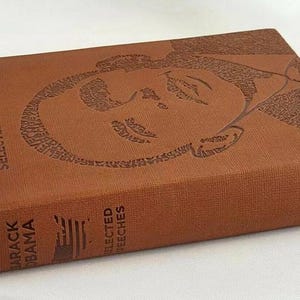 May include: A brown leather-bound book with the title "Barack Obama Selected Speeches" embossed on the cover. The cover features a portrait of Barack Obama created from the words of his speeches.
