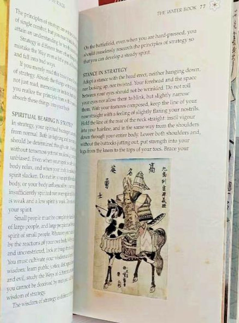 K&ouml;nnte beinhalten: Eine Schwarzwei&szlig;illustration eines Samurai-Kriegers zu Pferd. Der Krieger tr&auml;gt eine R&uuml;stung und einen Helm mit einem Wappen. Das Pferd tr&auml;gt einen Sattel und hat ein geflecktes Muster. Die Illustration ist von japanischem Text umgeben.