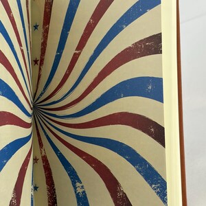 May include: Open notebook with pages displaying a vintage design of red, white, and blue stripes radiating from the center. Small red and blue stars are scattered across the cream-coloured background. The notebook has a brown cover.