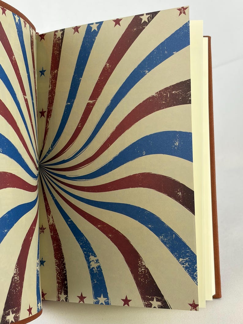 May include: Open notebook with pages displaying a vintage design of red, white, and blue stripes radiating from the center. Small red and blue stars are scattered across the cream-coloured background. The notebook has a brown cover.