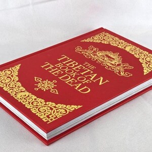 K&ouml;nnte beinhalten: Ein rotes Hardcover-Buch mit dem Titel "Das tibetische Totenbuch" mit goldenen Verzierungen. Der Einband zeigt verzierte goldene Designs in den Ecken und um den Titel. Das Buch liegt auf einer wei&szlig;en Oberfl&auml;che.