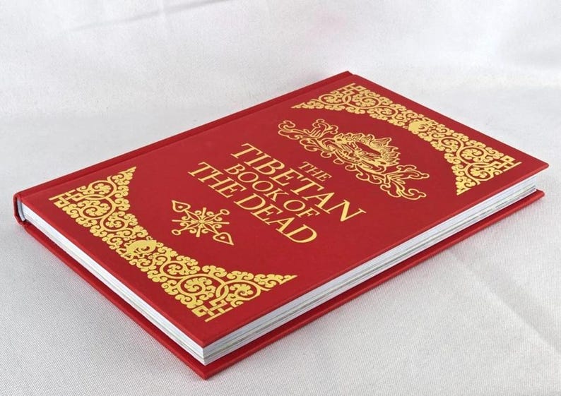 K&ouml;nnte beinhalten: Ein rotes Hardcover-Buch mit dem Titel "Das tibetische Totenbuch" mit goldenen Verzierungen. Der Einband zeigt verzierte goldene Designs in den Ecken und um den Titel. Das Buch liegt auf einer wei&szlig;en Oberfl&auml;che.