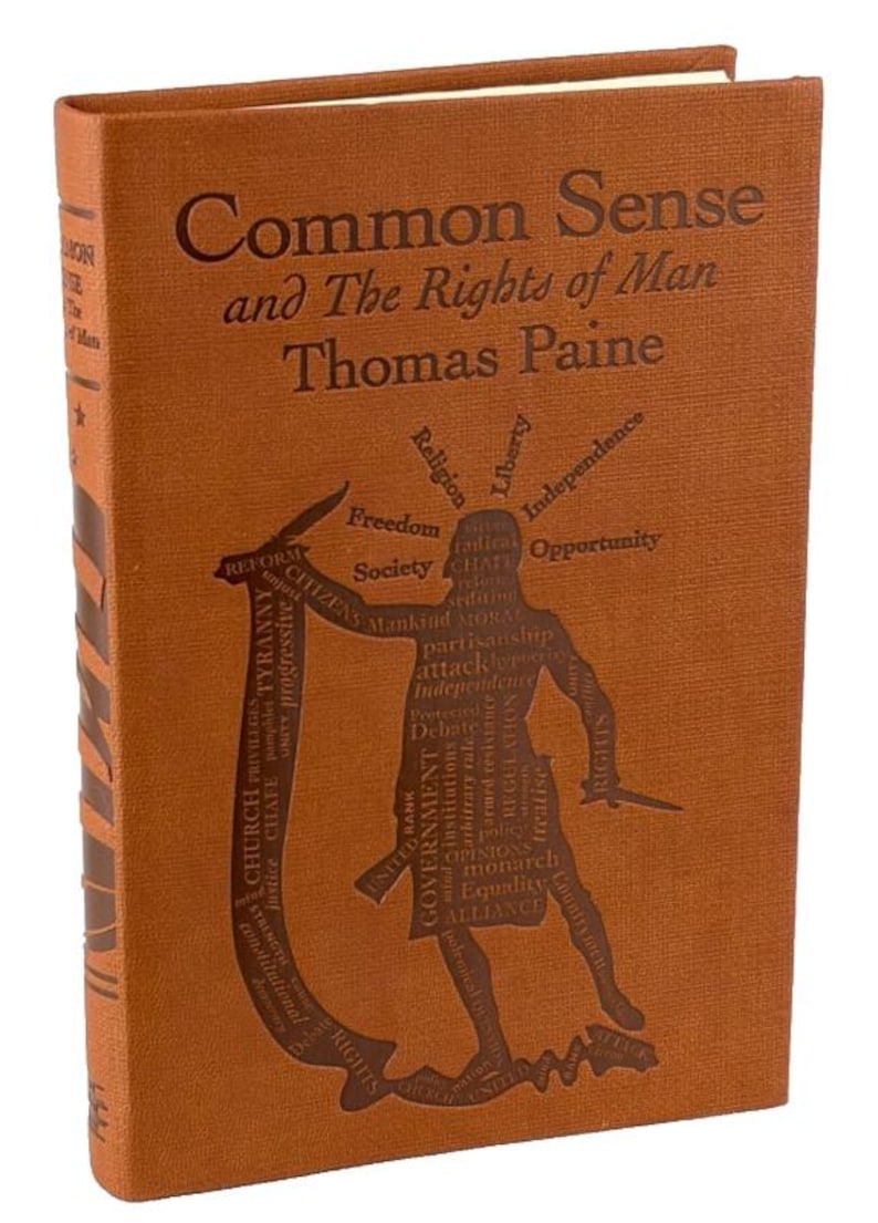 May include: A brown hardcover book titled "Common Sense and The Rights of Man" by Thomas Paine. The cover features embossed text and a graphic of a figure with knives, surrounded by words such as "Freedom" and "Government".