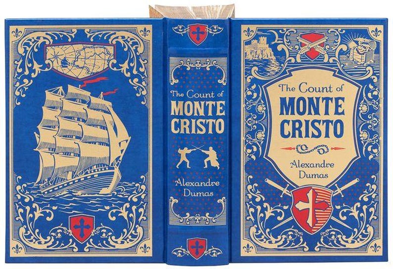 May include: A blue hardcover book with gold accents and a title that reads "The Count of Monte Cristo" by Alexandre Dumas. The cover features a ship sailing on the sea, two figures fencing, and a crest with a cross.