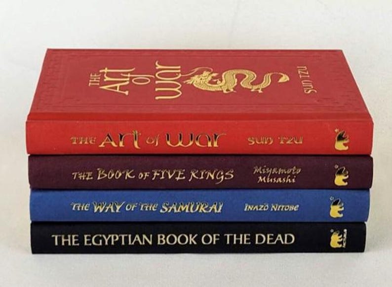 May include: A stack of five hardcover books with gold lettering on the spines. The titles are "The Art of War", "The Art of War", "The Book of Five Rings", "The Way of the Samurai", and "The Egyptian Book of the Dead".