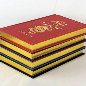 May include: A stack of four books with gold-gilded edges. The top book is red with a gold dragon and the title "The Art of War" by Sun Tzu.