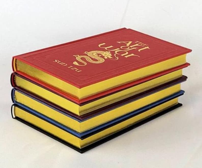 May include: A stack of four books with gold-gilded edges. The top book is red with a gold dragon and the title "The Art of War" by Sun Tzu.