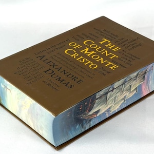 May include: A hardcover book titled "The Count of Monte Cristo" by Alexandre Dumas. The book has a brown cover with gold lettering and a detailed illustration on the edges. The book's pages are visible.