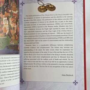 K&ouml;nnte beinhalten: Offenes Buch mit Text &uuml;ber Das tibetische Totenbuch, mit rotem Einband und dekorativem Design. Zwei kleine Messingbecken sind oben am Buch befestigt. Seite 15 ist sichtbar.