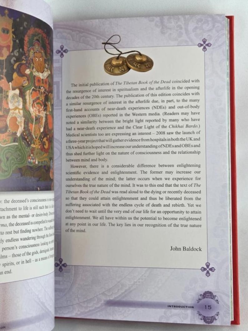 K&ouml;nnte beinhalten: Offenes Buch mit Text &uuml;ber Das tibetische Totenbuch, mit rotem Einband und dekorativem Design. Zwei kleine Messingbecken sind oben am Buch befestigt. Seite 15 ist sichtbar.