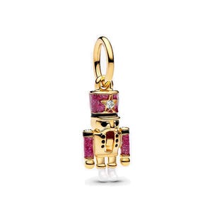 May include: A gold-colored charm in the shape of a nutcracker soldier. The charm features a red and pink glittery hat and arms, with a star-shaped accent. The soldier has white boots and black details. A small, clear gem is set in the star.