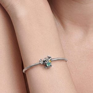 Op de afbeelding: Een zilveren slangenkettingarmband met een bedel in de vorm van een stripfiguur met grote oren en zwarte ogen. Het figuurtje houdt een klein groen voorwerp met een geel accent vast, gemaakt van gepolijst zilverkleurig metaal met details.