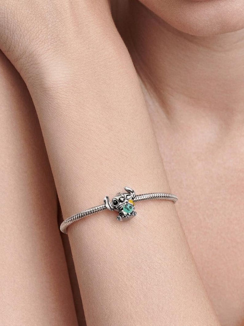 Op de afbeelding: Een zilveren slangenkettingarmband met een bedel in de vorm van een stripfiguur met grote oren en zwarte ogen. Het figuurtje houdt een klein groen voorwerp met een geel accent vast, gemaakt van gepolijst zilverkleurig metaal met details.