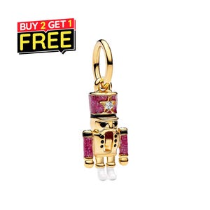May include: A gold-toned nutcracker charm featuring a glittery magenta hat and sleeves, black accents, and white boots. The hat is adorned with a small, clear, sparkling stone. The charm includes a circular gold jump ring at the top for attachment.