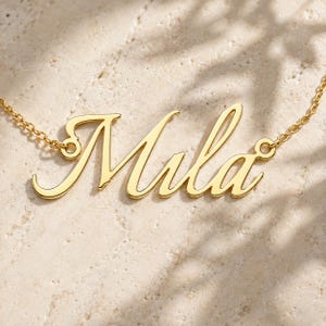 May include: A gold-toned metal necklace featuring a nameplate pendant with the name Mila written in a cursive script font. The pendant is attached to a delicate gold-toned chain on both sides, showcasing a polished finish and smooth curves.
