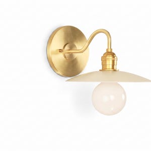 May include: A brass wall sconce with a circular backplate and curved arm. The sconce features a cream-colored shade and a round, white light bulb. The fixture is designed for indoor lighting.