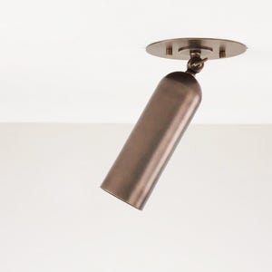 Puede incluir: Foco de techo cilíndrico de color bronce, montado en un techo blanco. La luz está orientada hacia abajo, fijada a una base circular con tres tornillos. La luminaria es de diseño moderno.