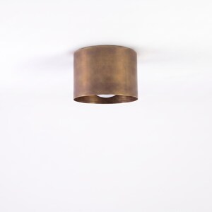May include: A cylindrical ceiling light fixture with a bronze finish. The light is mounted flush against a white ceiling. The fixture has a simple, modern design, and the light source is visible at the bottom.