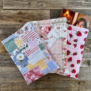 Könnte beinhalten: Drei gesteppte Buchhüllen mit Blumen- und Fruchtmustern. Eine Hülle zeigt Erdbeeren auf rosa Grund, eine andere Rosen und die dritte ein Patchwork-Design. Jede Hülle hat ein Etikett mit der Aufschrift "Cedar Hill Studios".